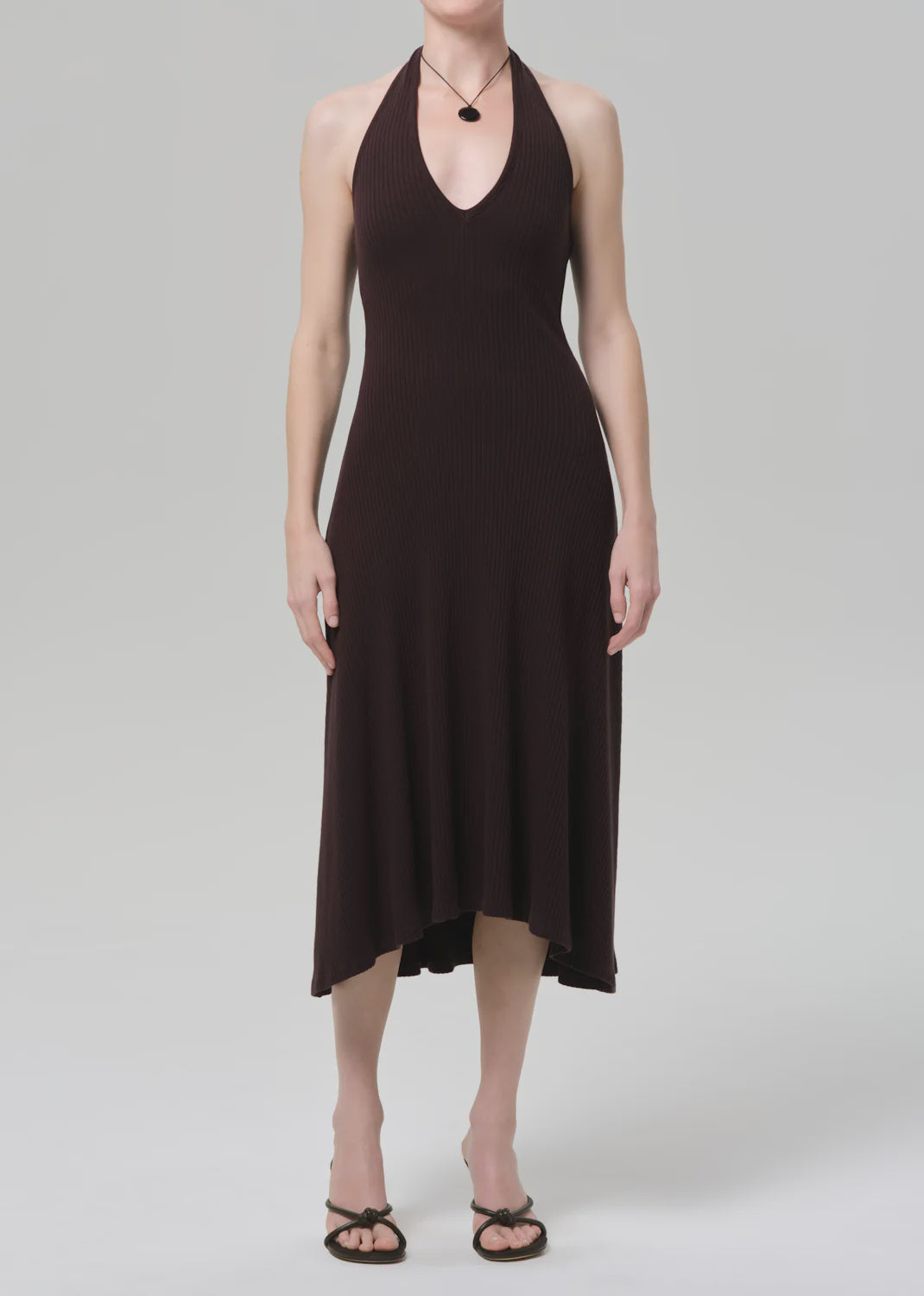 A person wears the mila halter dress by Citizens Of Humanity/AGOLDE in chocolate brown with a subtle ribbed texture, paired with black strappy sandals, standing against a plain light gray background.