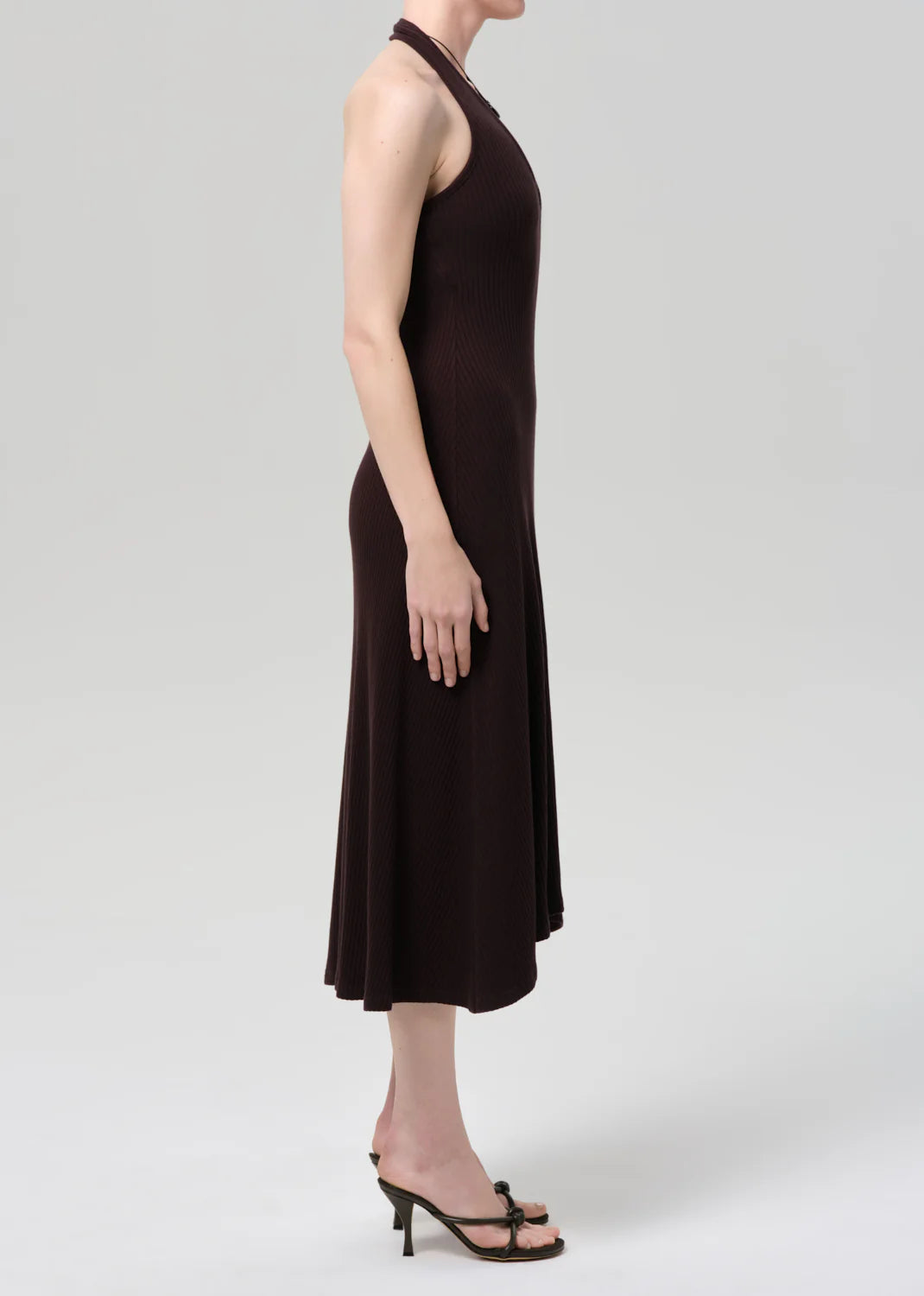 A person stands in profile wearing the Citizens Of Humanity/AGOLDE mila halter dress in chocolate brown with black heeled sandals against a plain light gray background.