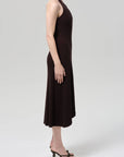 A person stands in profile wearing the Citizens Of Humanity/AGOLDE mila halter dress in chocolate brown with black heeled sandals against a plain light gray background.