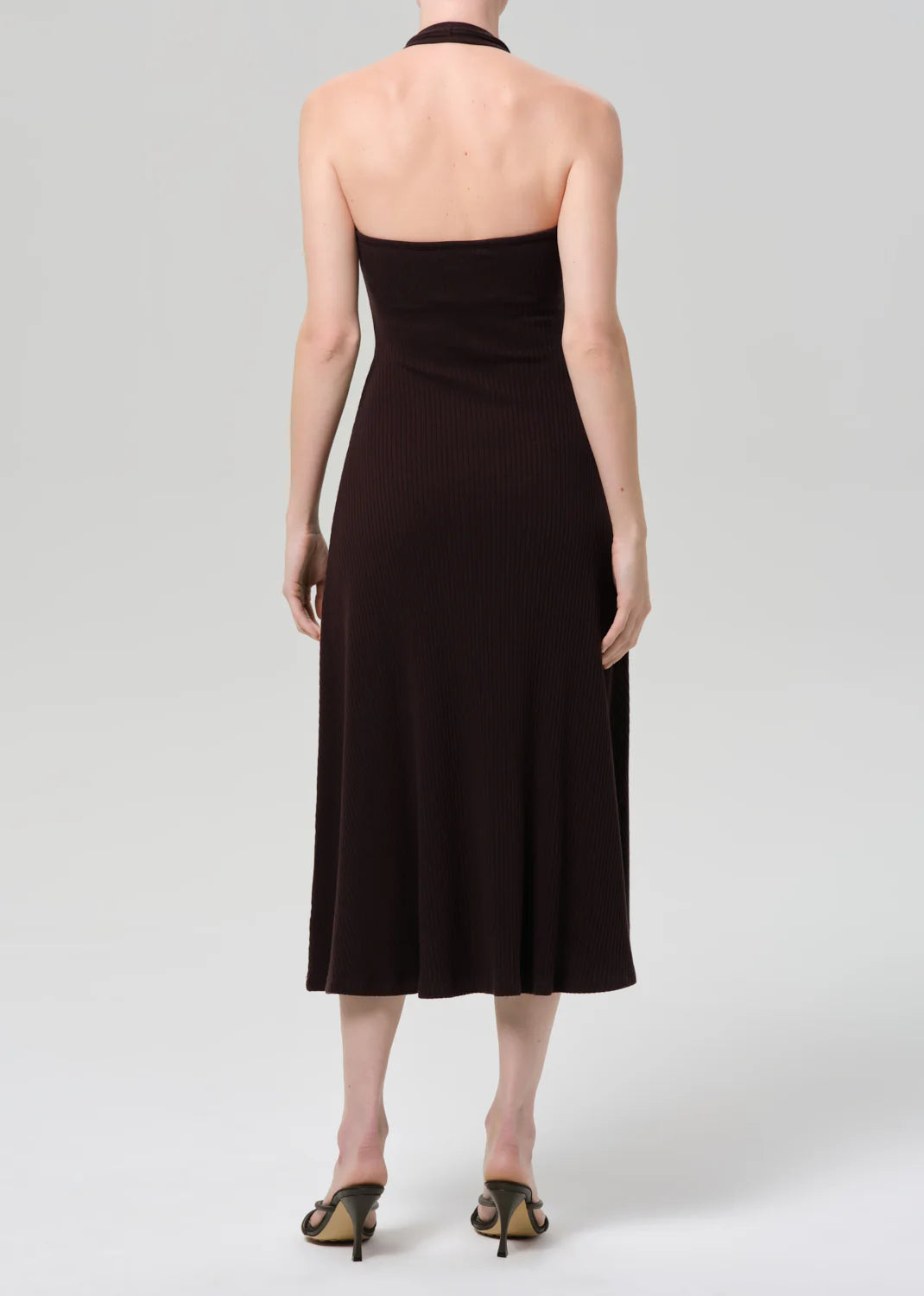 A person is seen from behind wearing the Citizens Of Humanity/AGOLDE mila halter dress in chocolate brown with heeled sandals, standing against a plain light gray background.