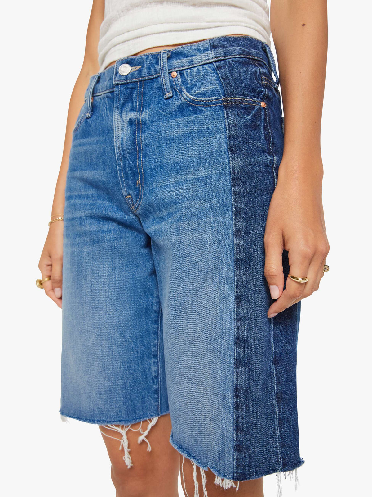 A person wears the Mother Mid Rise Maven Bermuda Fray shorts featuring a light blue front, dark blue sides, raw frayed hems, metal button and pocket details, paired with a white ribbed tank top for a stylish wide leg look.