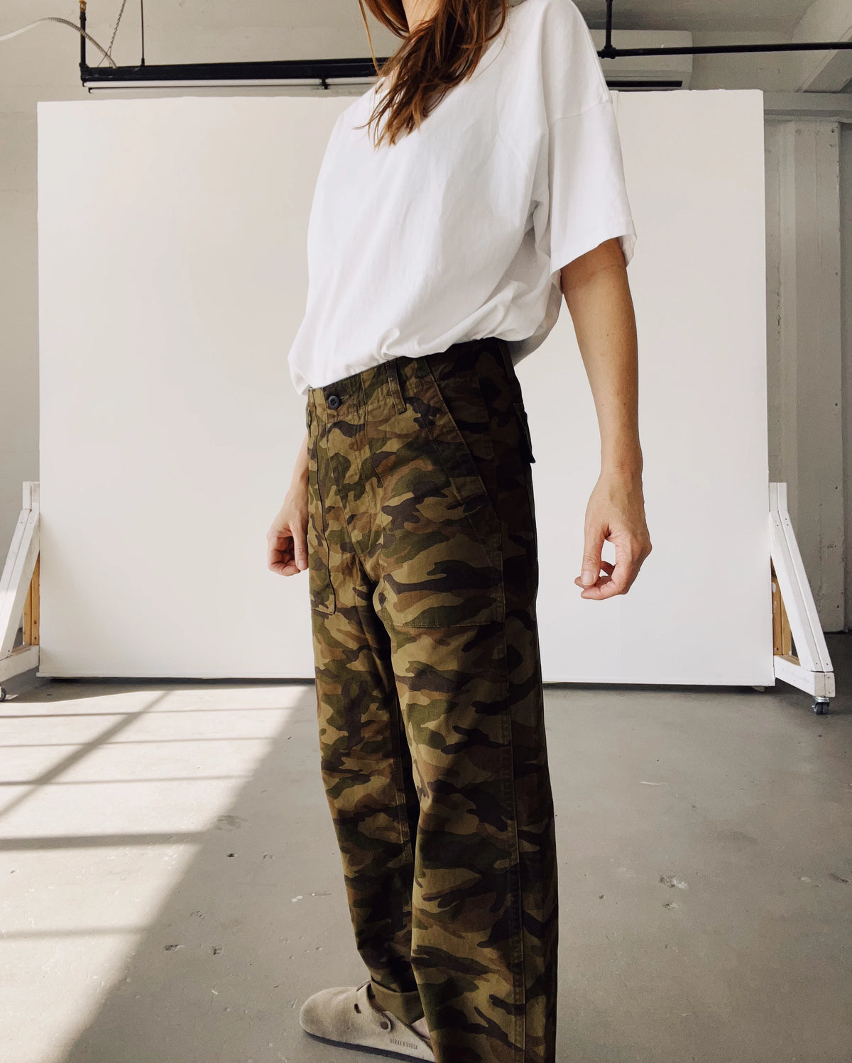 Indoors, a person wears Hey Gang's The Officer Pant in cotton ripstop camouflage, paired with a loose white t-shirt and beige shoes against a minimal white background. The person's face is not shown.