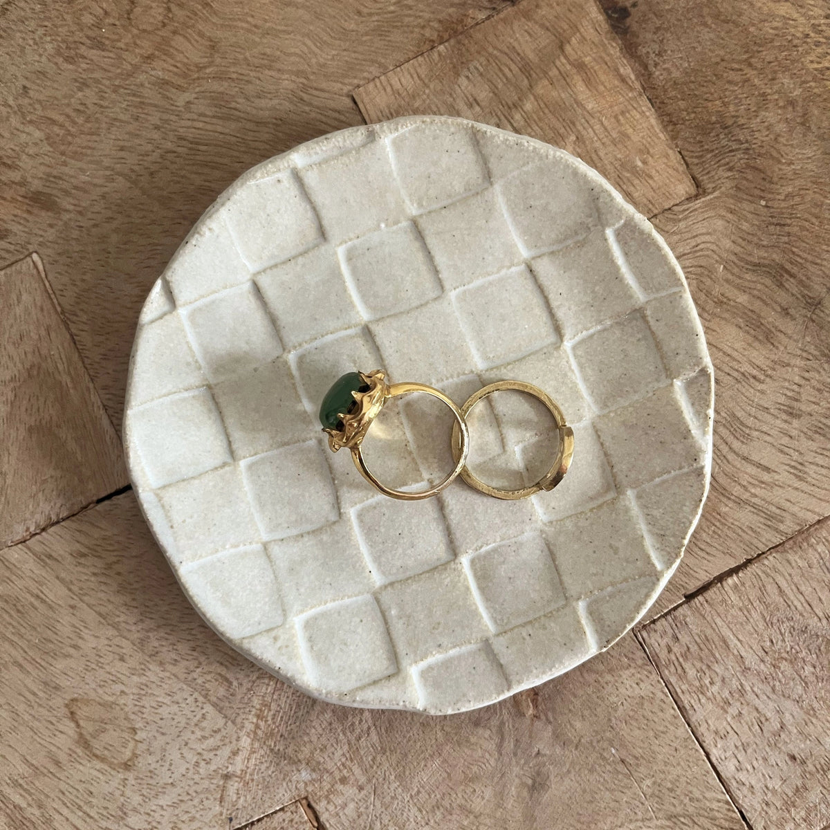 Two gold rings, one with a green stone, rest on Faire’s Ghost Checker Trinket Dish—a round, off-white glazed stoneware dish with a subtle checker pattern—displayed on a wooden surface.