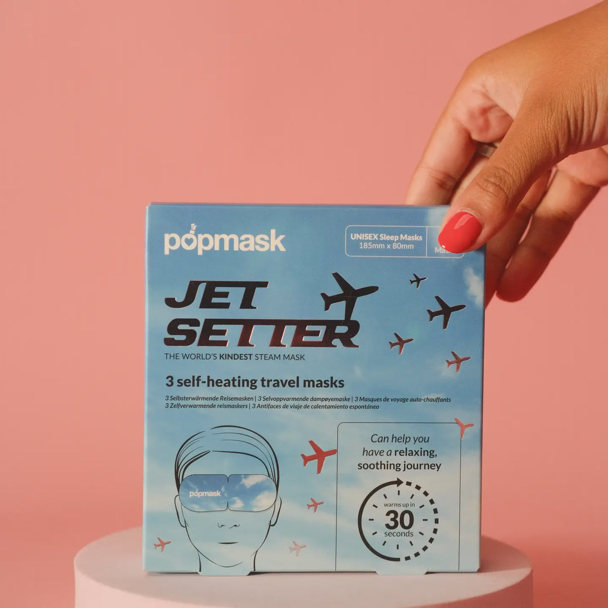 A hand with pink nail polish holds a box of "Faire Eye Masks" against a pink background. The packaging showcases sky and plane graphics, promoting the unisex steam masks as ideal for a relaxing journey within 30 seconds.