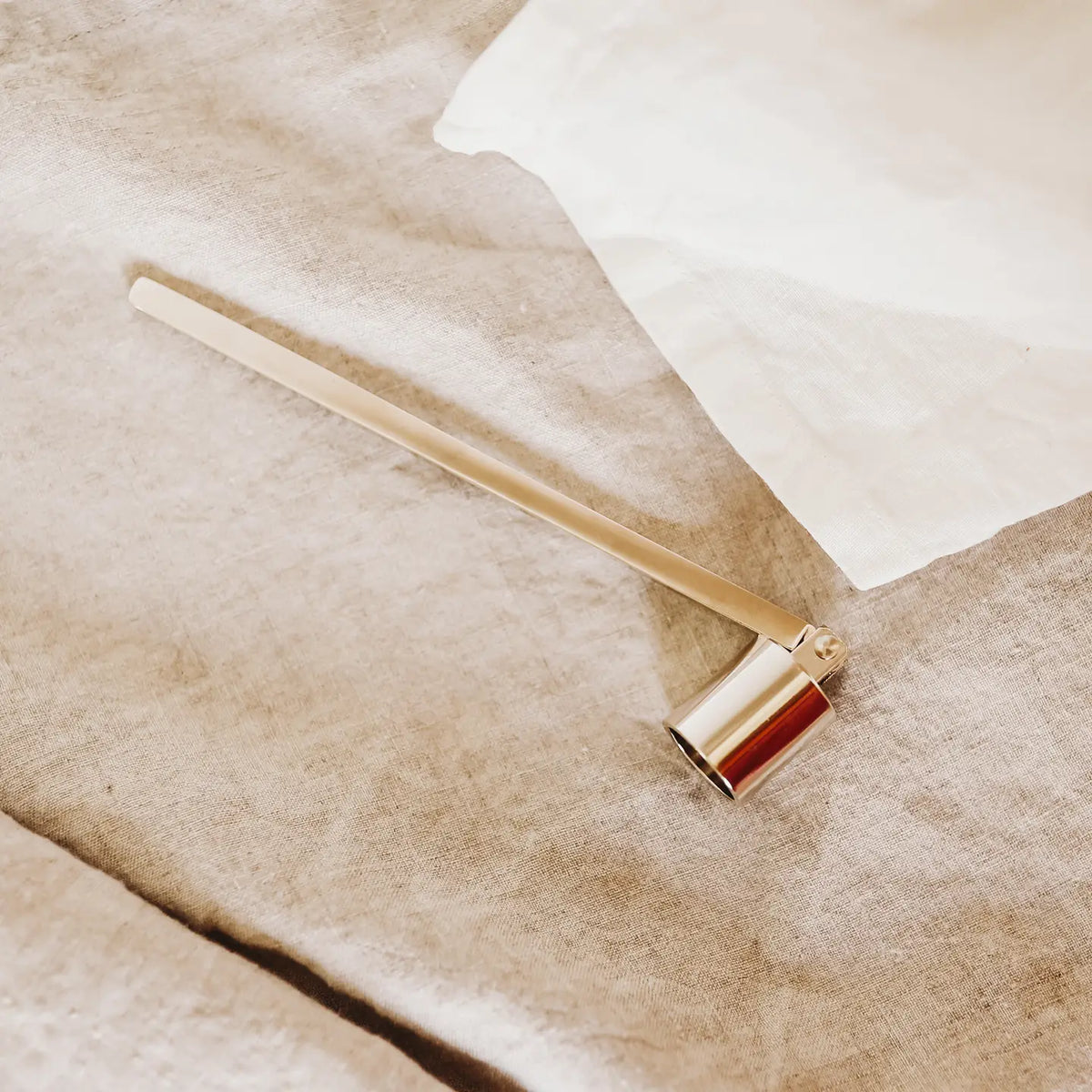 The Faire Candle Snuffer, a sophisticated candle accessory, rests on a beige linen surface beside a white cloth.