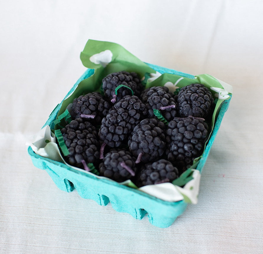 A teal carton with a green paper lining holds Faire’s Blackberry Beeswax Candles, inspired by the look of fresh blackberries, and sits on a light-colored surface.