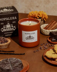 A lit Faire Nopalera Chocolate Candle sits on a table with cinnamon sticks, chocolate, vanilla beans, cookies, cacao nibs, and a marigold, evoking spiced hot chocolate. The candle’s box appears in the background.