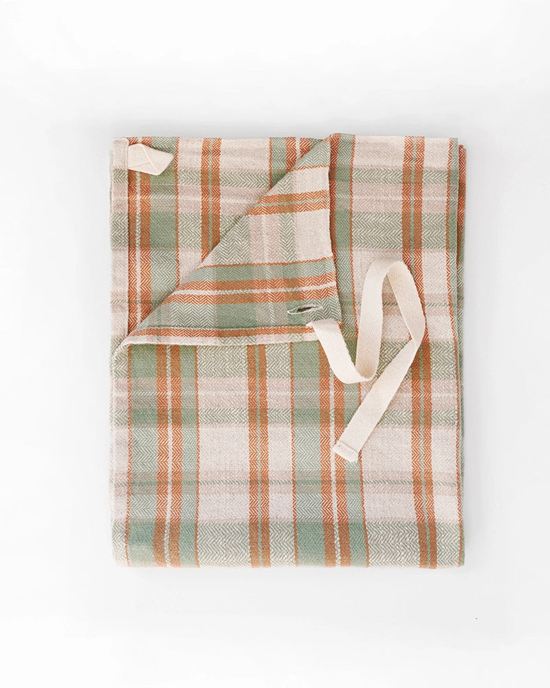 The Faire Cook's Cloth is a folded apron in a soft cotton-linen blend with green, beige, and orange plaid stripes and cream waist ties, shown on a white background.