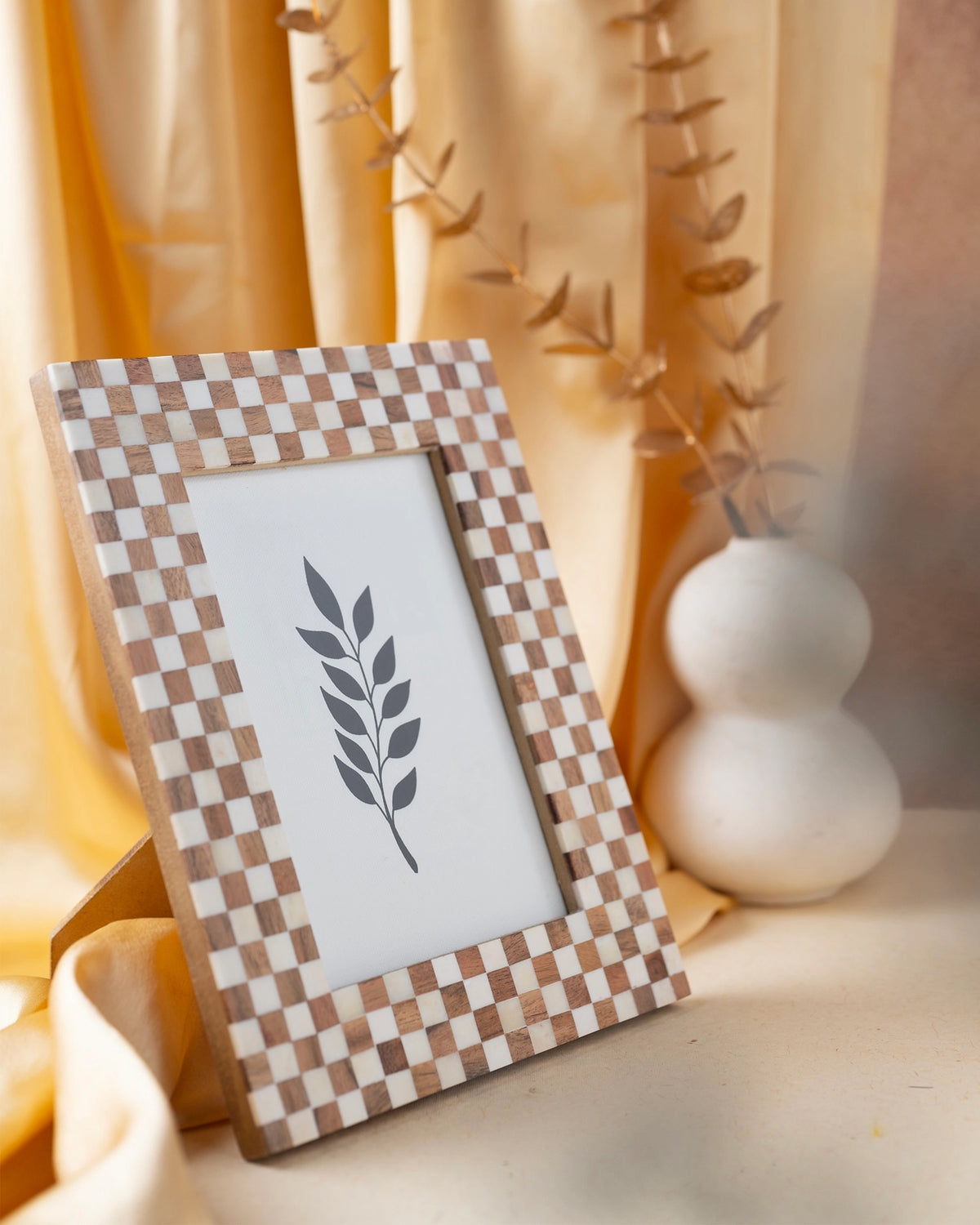 Bone and wood checkered 4x6 frame by A Faire displayed on a yellow cloth beside a white vase with dried branches.