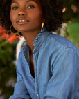 A woman with curly hair and large hoop earrings poses outdoors among green and red foliage, wearing the Frank & Eileen Mary Classic Shirtdress- Ruffle.