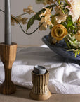 A close-up of a table featuring the Accent Decor Argon Candleholder in brown holding a gray candle, alongside a ceramic candleholder with a melted candle and a floral arrangement in a blue vase on white cloth.