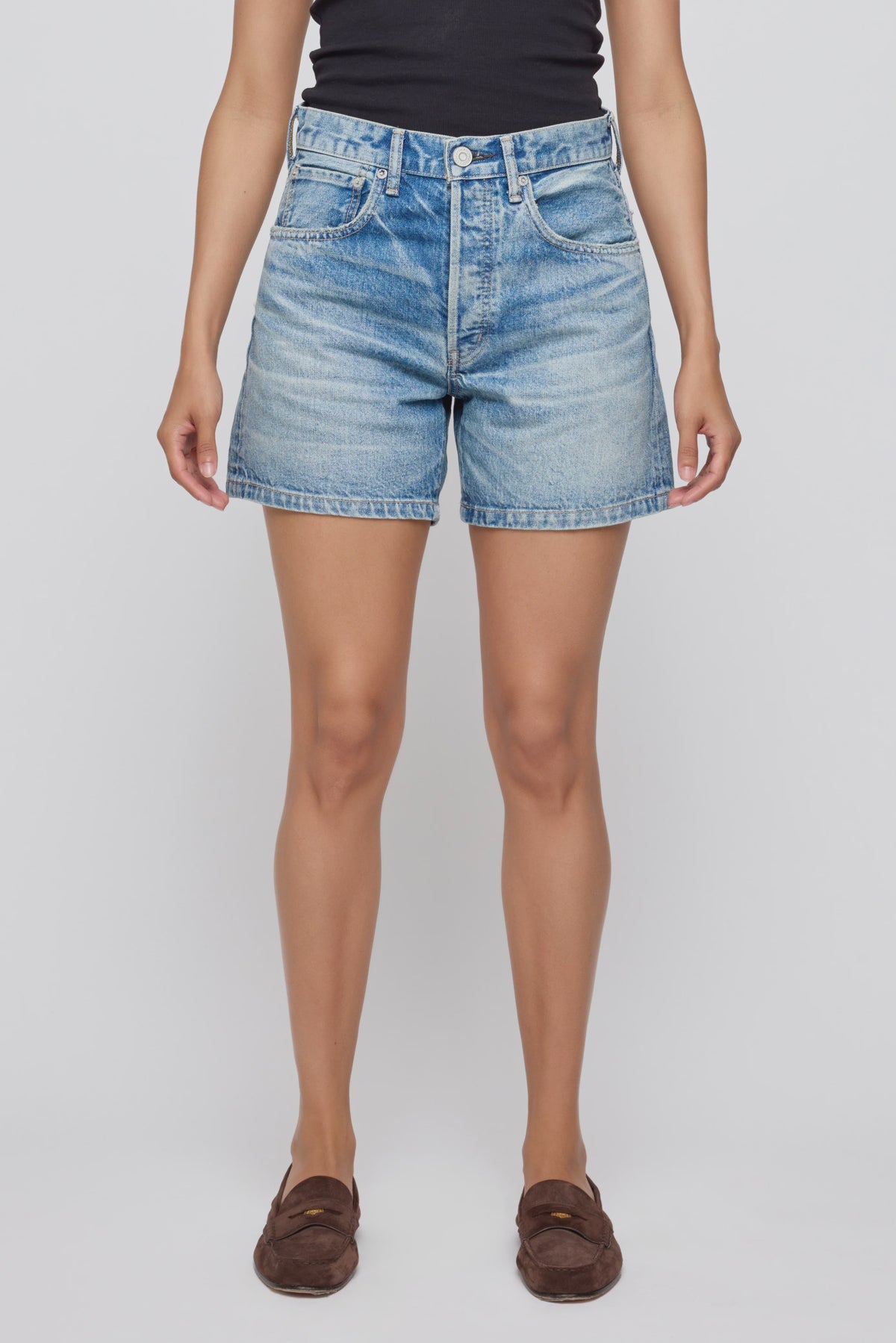 A person wearing a black sleeveless top, brown loafers, and Moussy’s Harrisburg Long Shorts—light blue high-waisted button fly jean shorts—stands against a plain white background.