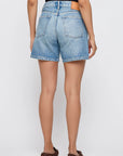 A person wearing a black top, brown shoes, and Moussy Harrisburg Long Shorts (light stretch, mid rise, button fly) is standing facing away from the camera against a plain light background.