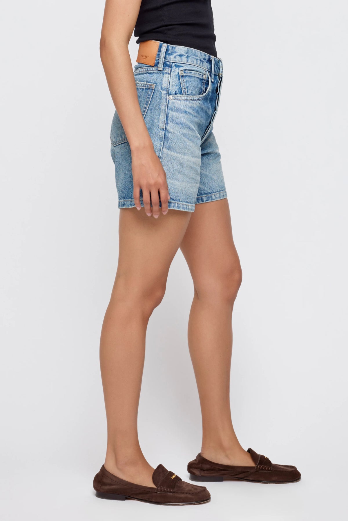 A person stands sideways against a plain white background, wearing Moussy Harrisburg Long Shorts in light blue stretch denim with a button fly, paired with a black top and brown loafers.