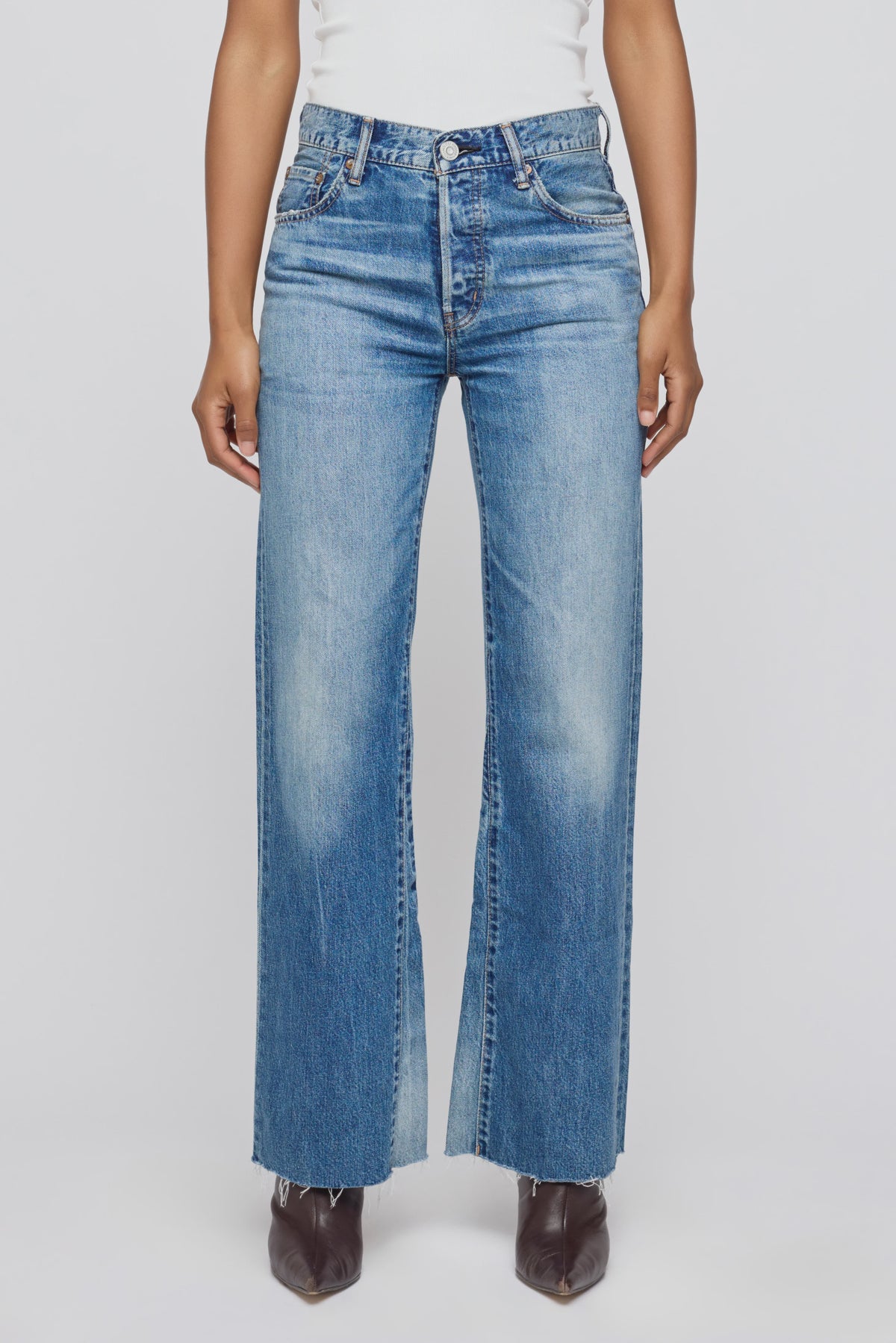 A person wearing Moussy Asheville Remake Flare high-waisted, light blue wide-leg jeans with a button fly and raw hem, paired with a white top and brown pointed-toe shoes, shown waist down against a plain background.