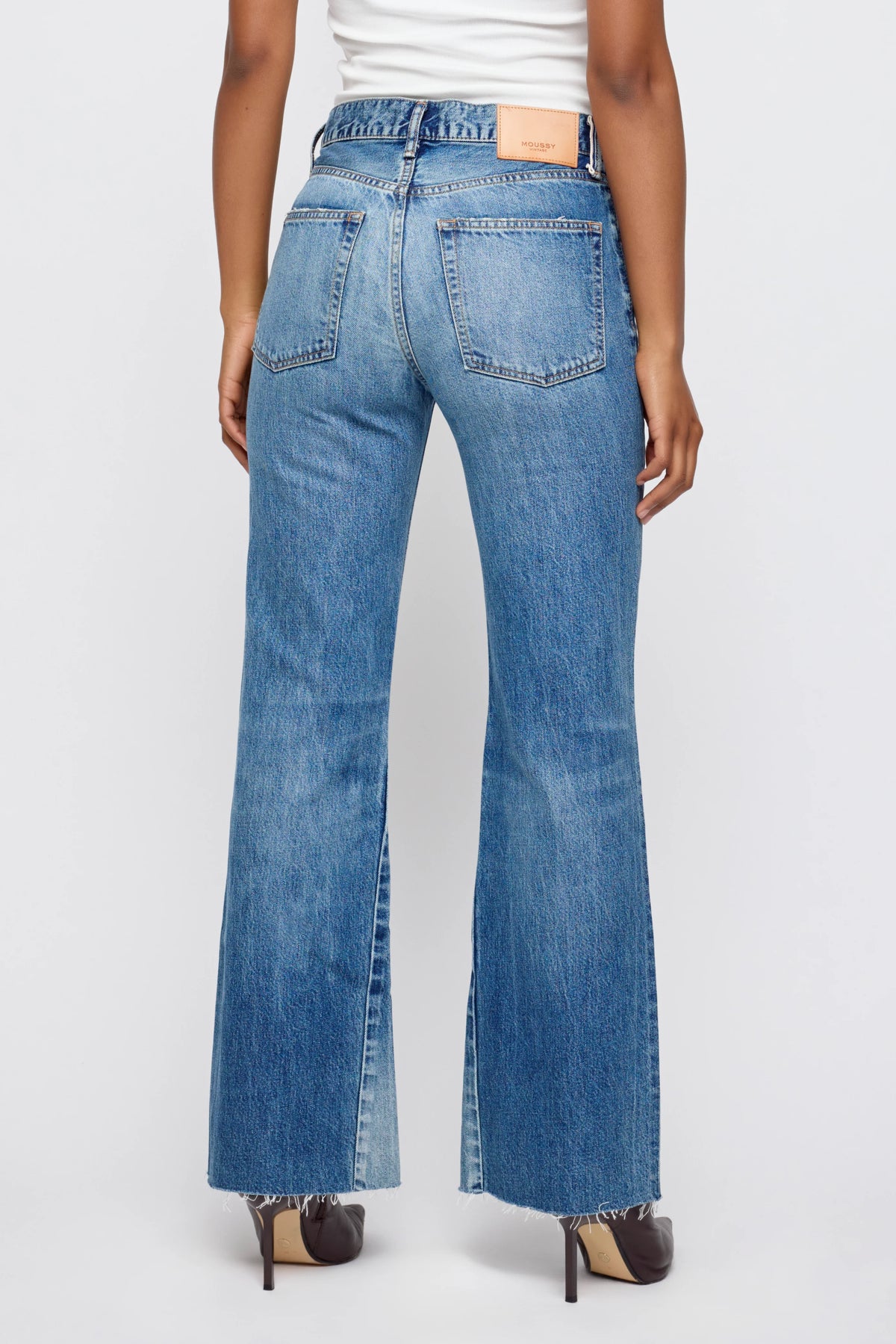 Someone is seen from behind wearing Moussy Asheville Remake Flare high-waisted blue jeans with frayed hems and front slits, paired with a white top and brown-heeled shoes, standing against a plain light background.