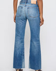 Someone is seen from behind wearing Moussy Asheville Remake Flare high-waisted blue jeans with frayed hems and front slits, paired with a white top and brown-heeled shoes, standing against a plain light background.