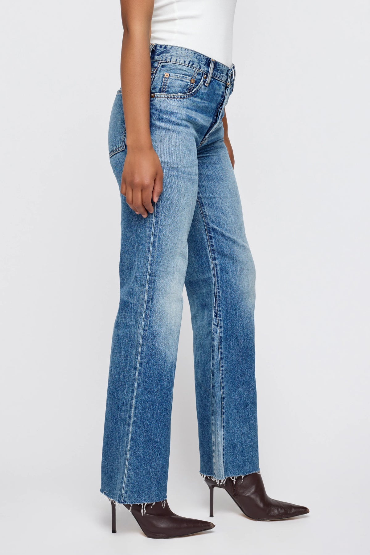 Wearing Moussy's Asheville Remake Flare jeans—lightweight blue denim with a raw hem—styled with a white top and pointed brown heeled boots, standing against a plain light background.