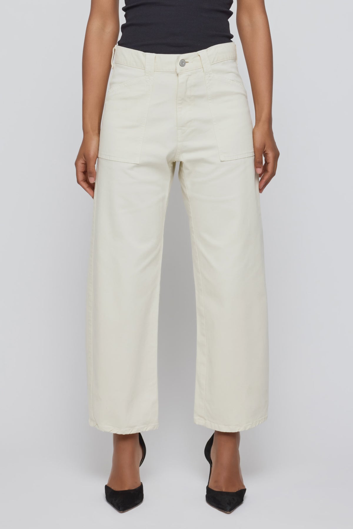 A person stands against a plain light background wearing Moussy Pomonok Gusset Cargo jeans in off-white with front patch pockets, paired with a black top and black pointed-toe heels.