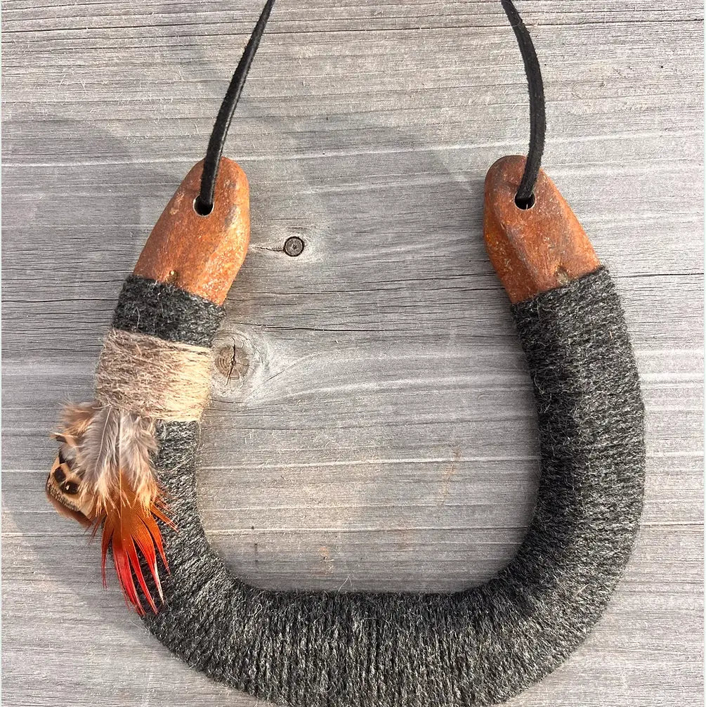 The Faire Lucky Horseshoe features an upcycled horseshoe wrapped in gray wool with wooden ends, twine, and feathers, hanging by a black cord—making it a unique housewarming gift.