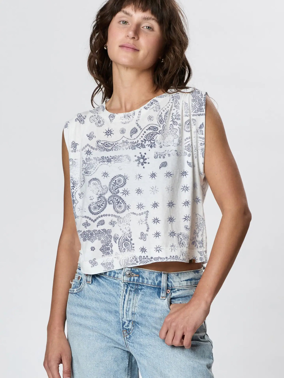 A person models Faire’s Hemp Jackie Pleat Shell Bandana, a sleeveless white top featuring a blue paisley and star print, styled with light blue jeans and standing against a plain light background.