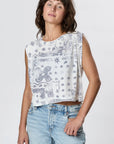 A person models Faire’s Hemp Jackie Pleat Shell Bandana, a sleeveless white top featuring a blue paisley and star print, styled with light blue jeans and standing against a plain light background.