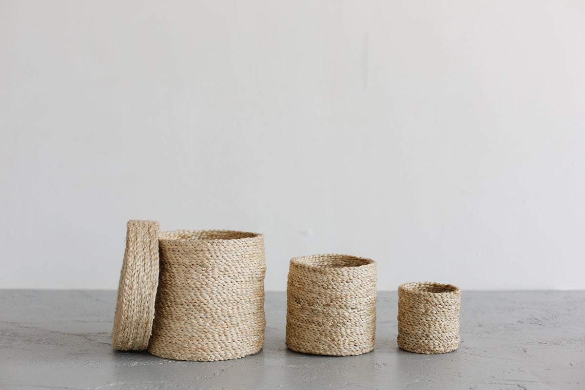 Three Faire Round Lidded Jute Baskets in different sizes are lined up on a gray surface with a plain white background. The largest basket's lid is propped against it.