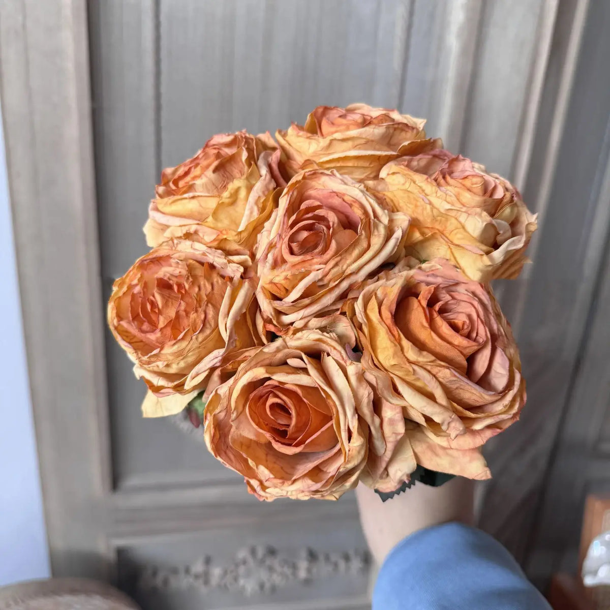 A hand holds the Faire 17'' Faux Vintage Rose Bouquet with curled petals, set against a light wooden background featuring carved panel details.