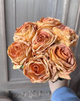 A hand holds the Faire 17'' Faux Vintage Rose Bouquet with curled petals, set against a light wooden background featuring carved panel details.