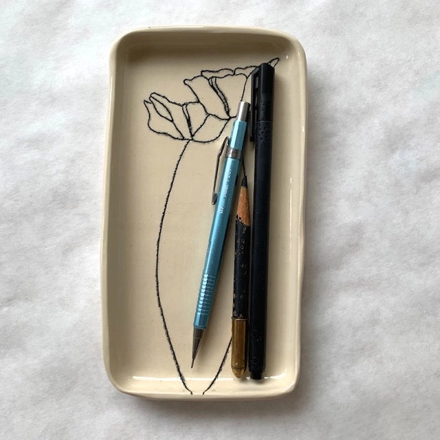 The Faire Floral Small Tray, featuring a hand-etched black flower outline on ceramic, holds a blue mechanical pencil, black pen, and short wooden pencil on a white surface.