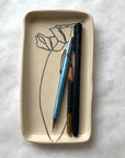 The Faire Floral Small Tray, featuring a hand-etched black flower outline on ceramic, holds a blue mechanical pencil, black pen, and short wooden pencil on a white surface.