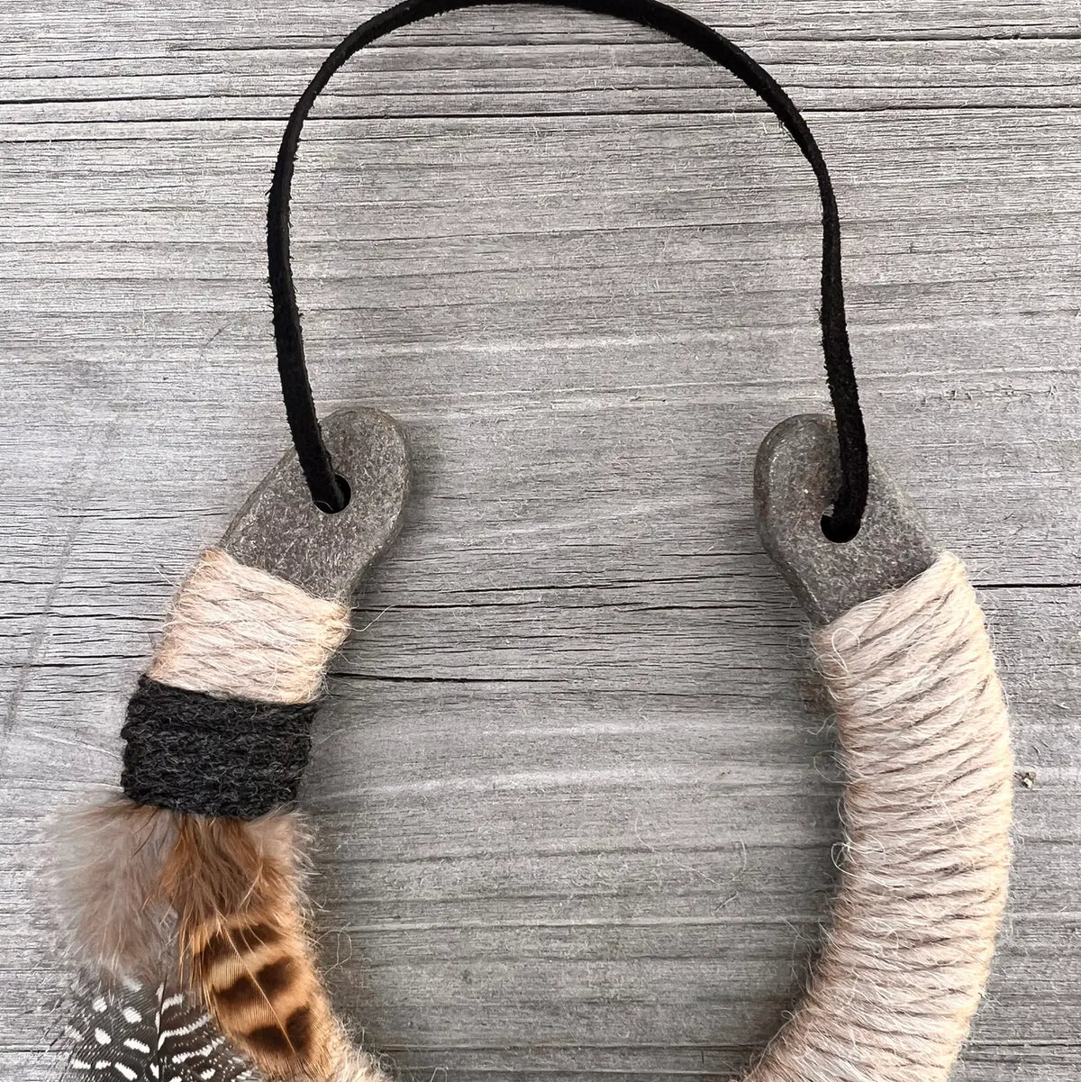 The Lucky Horseshoe by Faire, wrapped in beige and gray twine with left-side feathers and a black cord, sits on weathered wood—perfect as a personalized or unique housewarming gift.