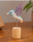 Purple hand-painted carved hummingbird by Faire on a wooden stand, displayed on a brown table.