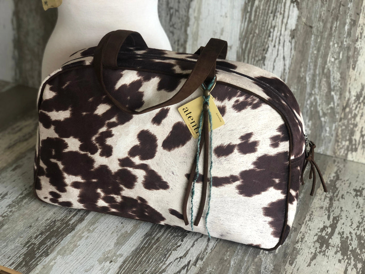 The Faire Oakley Overnighter—Faux Cowhide features a brown and white cowhide pattern, dark brown handles, and a yellow tag with blue and brown string. This vegan weekender bag is perfect for your next overnight getaway.
