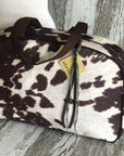 The Faire Oakley Overnighter—Faux Cowhide features a brown and white cowhide pattern, dark brown handles, and a yellow tag with blue and brown string. This vegan weekender bag is perfect for your next overnight getaway.
