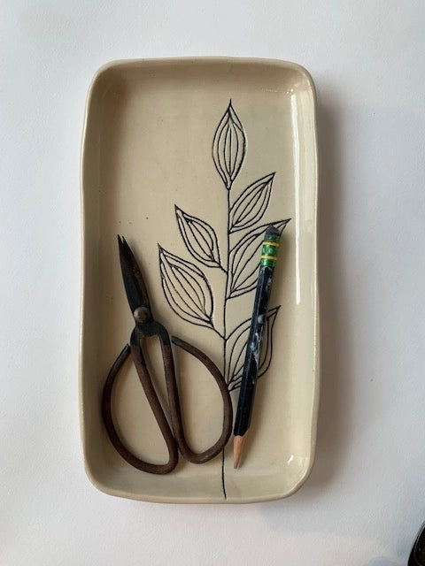 The Floral Small Tray by Faire, a rectangular hand-etched ceramic piece with a black leaf design, holds vintage scissors and a green-eraser pencil on a white surface, capturing subtle botanical symbolism.