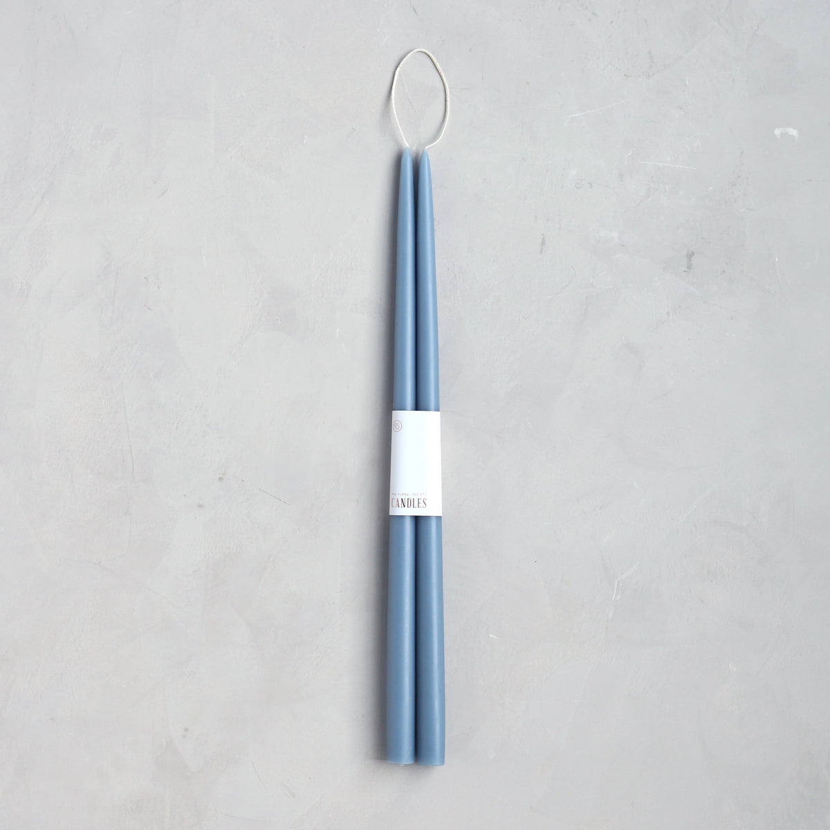 The Floral Society 18" Taper Candle Pair in blue features tall, slender candles bound with a white band and topped with a looped wick—an elegant touch for any home décor. Set on a light gray textured surface.