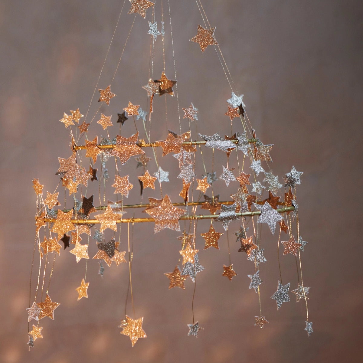 The Faire Gold Sparkle Star Chandelier features gold and silver glittery stars hanging from dual golden frames, softly lit on a neutral brown backdrop for an elegant, versatile accent in any space.
