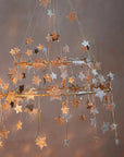 The Faire Gold Sparkle Star Chandelier features gold and silver glittery stars hanging from dual golden frames, softly lit on a neutral brown backdrop for an elegant, versatile accent in any space.