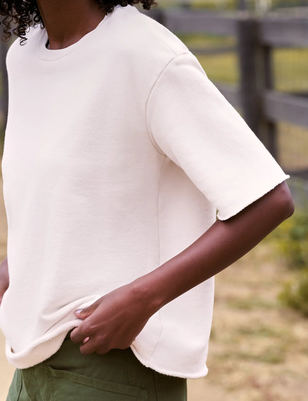 Woman in a casual crew neck tee by Frank & Eileen with green pants stands outdoors near a rustic wooden fence.