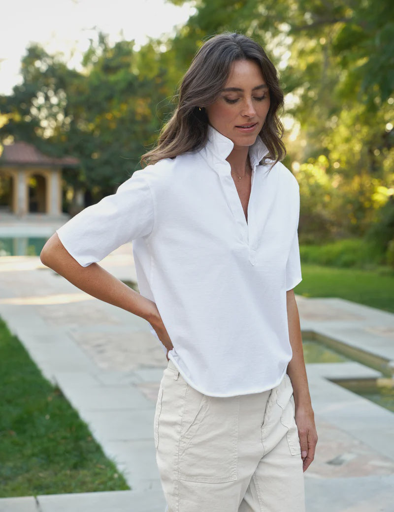 A woman with wavy brown hair stands outdoors on a stone pathway, wearing the Frank & Eileen Patty Popover SS Henley in white and light pants, with greenery and a pool in the background.