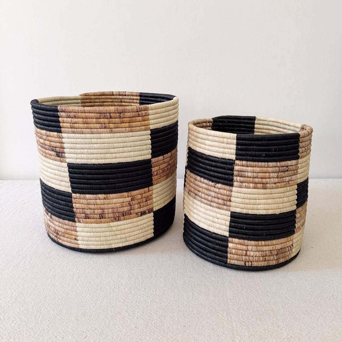Two handmade Faire Storage Plant Baskets: Zindiro, featuring a black, cream, and brown checkered pattern, are displayed on a light textured surface with a plain white background.