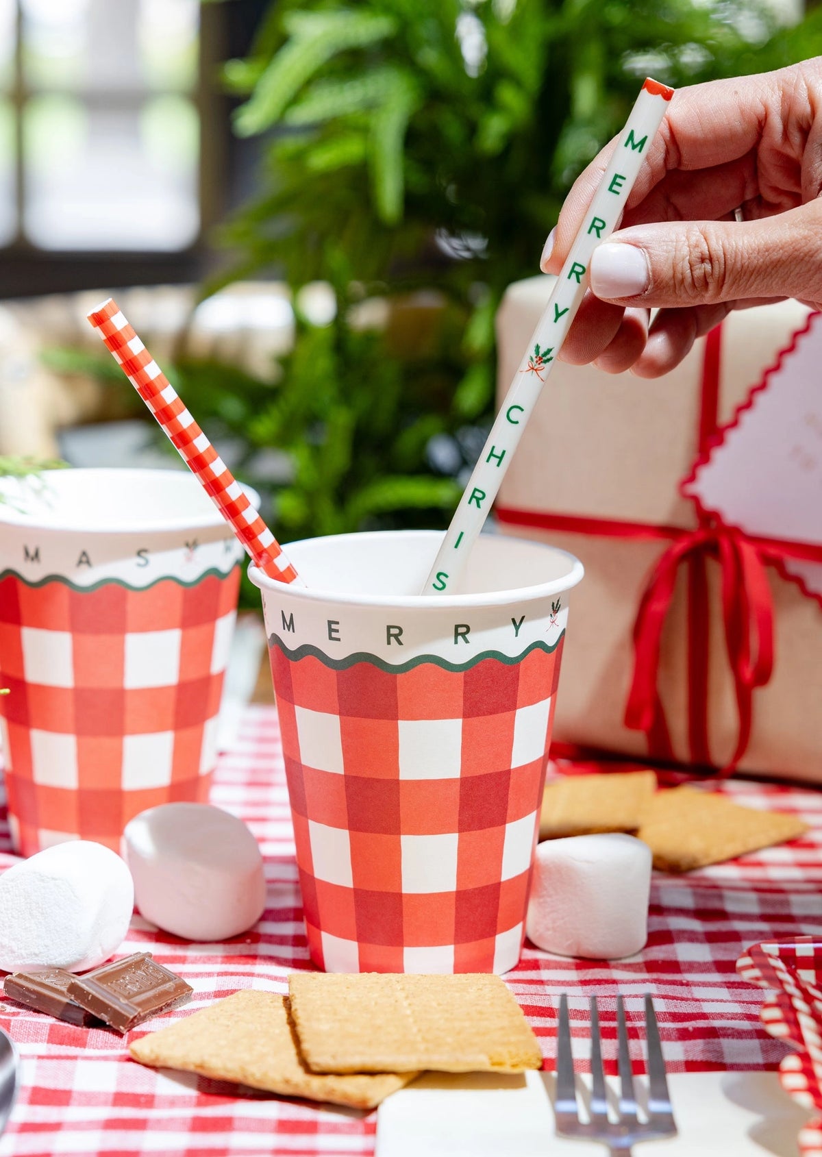 Red Gingham Merry Christmas Reusable Straw held over matching red gingham cups on a festive checkered tablecloth.
