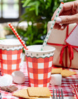 Red Gingham Merry Christmas Reusable Straw held over matching red gingham cups on a festive checkered tablecloth.