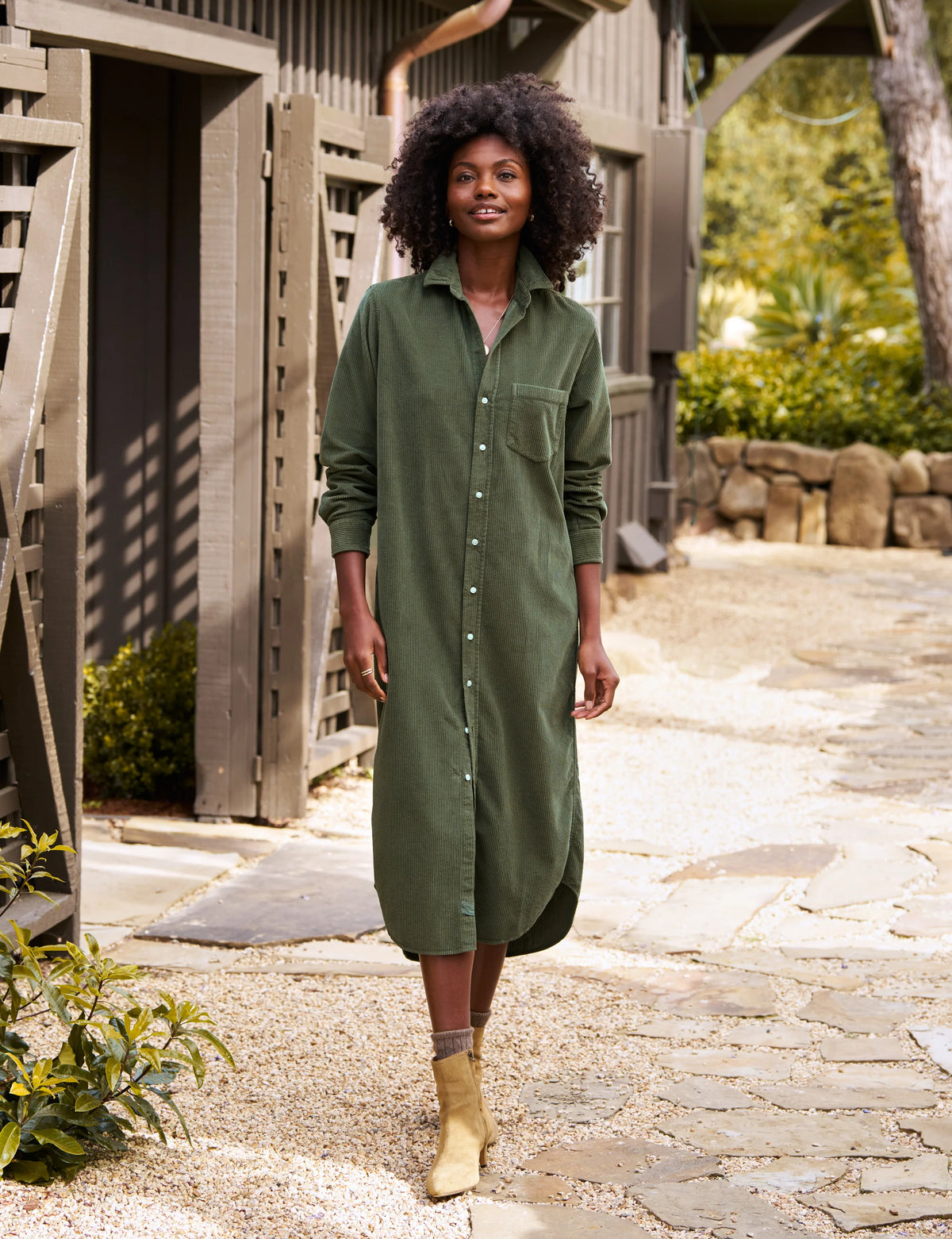 A woman with curly hair walks outdoors, smiling in the Frank & Eileen RORY Maxi Shirtdress and beige ankle boots. The rustic wooden building and greenery create a relaxed, stylish California-chic vibe.