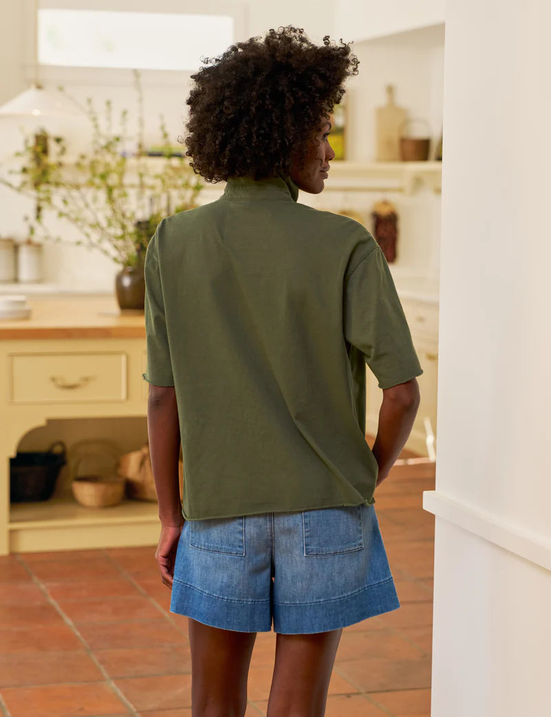 A person with short curly hair, wearing a Frank &amp; Eileen Patty Popover SS Henley in olive green and denim shorts, stands in a bright kitchen with tile floors and light-colored cabinets, facing away from the camera.