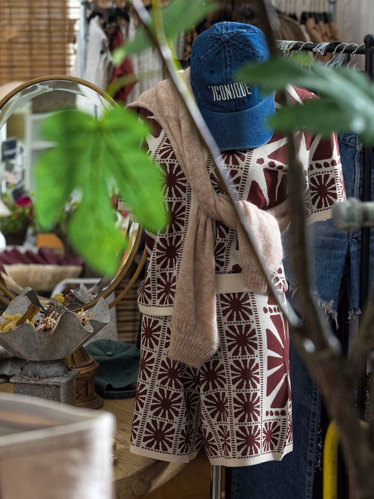Patterned romper with beige sweater and blue cap displayed on mannequin among plants at Bungalow boutique.