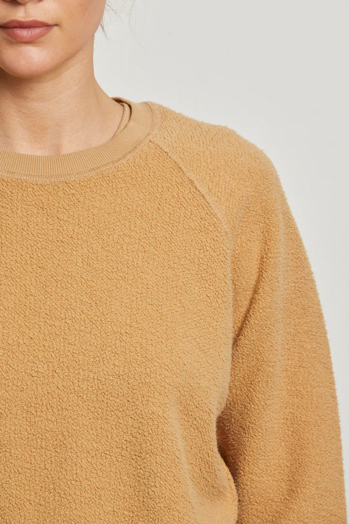 A close-up shows a person wearing the Perfectwhitetee Ziggy Reverse LS Sweatshirt in textured tan cotton fleece with a crew neckline, photographed from the shoulders up against a plain light background. Only the lower half of their face is visible.