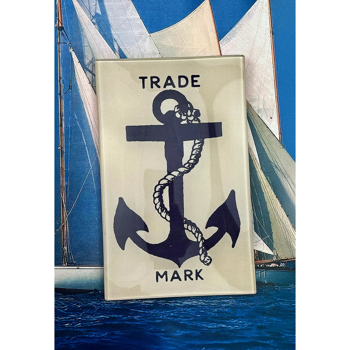 The Faire Shipman's Anchor Glass Tray showcases reverse decoupage artistry with a black anchor and rope labeled "TRADE MARK," displayed before a sailboat photo with white sails and blue sky.