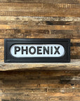 The Faire Lighted Sign features bold black “PHOENIX” lettering on a white background, displayed on a rustic wooden shelf against plank walls, capturing the vintage charm of a classic marquee.